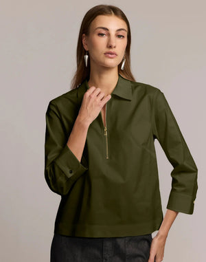 A woman with long brown hair wears the HINSON WU - ALESSA 3/4 SLEEVE SHIRT in olive green, featuring a zip-up collar and stretch polished cotton blend, paired with dark pants. She stands against a light background with minimal makeup and geometric earrings.