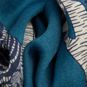 A blonde woman in a black coat stands against a white background, wearing the INOUI EDITIONS "Owls on a Branch" wool scarf in blue by INOUI, draped over her neck and one shoulder with intricate white owl designs.