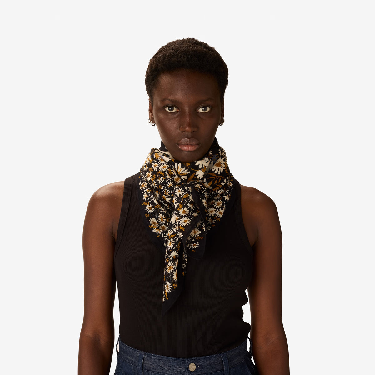 A woman with short hair, in a black sleeveless top and blue pants, stands against a white background. She wears the INOUI EDITIONS FAUST SCARF IN BLACK from INOUI, featuring yellow and white flowers, wrapped around her neck.
