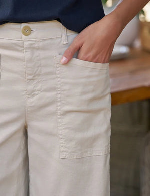 A person wearing the FRANK & EILEEN Limerick Cropped Wide Leg Carpenter Pant in Italian Performance Linen Cement, styled with tan lace-up shoes, stands indoors on a wooden floor with wicker furniture in the background.