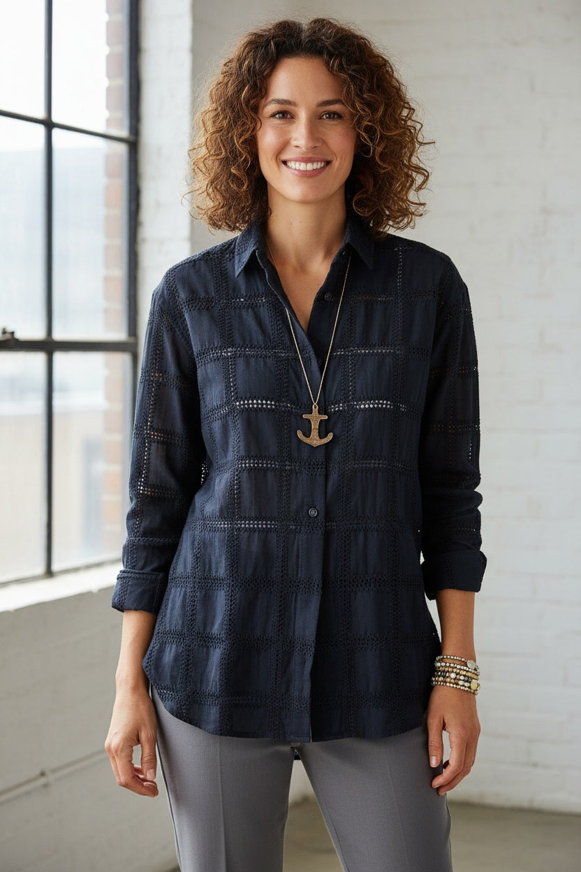 A person wearing the MERSEA Bronte Button Up Shirt in Navy Crochet holds a large bird-shaped pendant necklace. Only the torso and part of the face are visible, with one hand resting on the chest. The individual also wears colorful beaded bracelets.