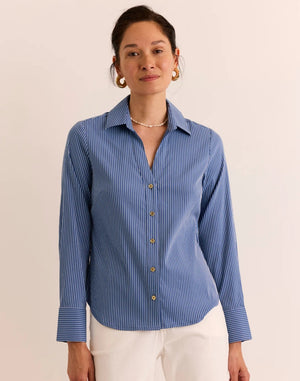 A woman stands against a plain, light background, exuding effortless style and breathable comfort in the HINSON WU Lindsey Long Sleeve Ribbon Stripe Shirt in Blue and White—a lightweight pinstripe button-up featuring gold buttons—paired with white pants.