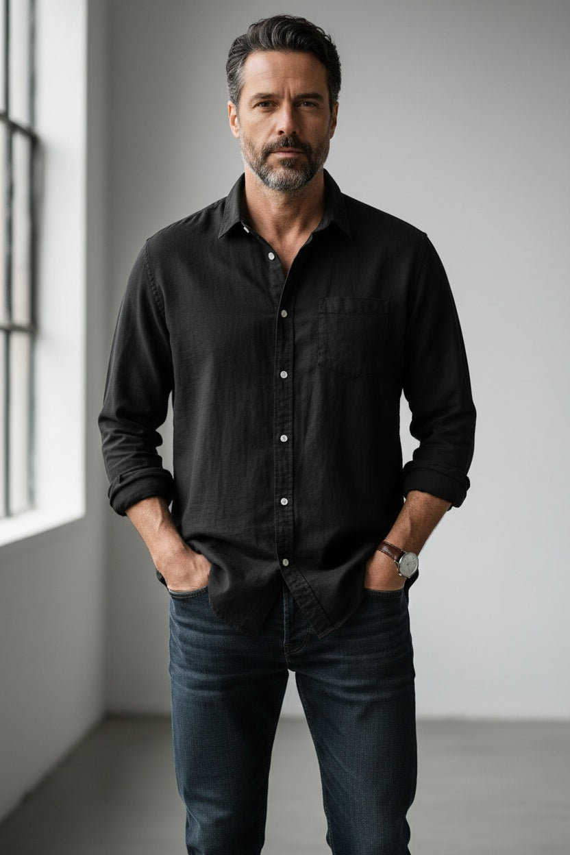 A man with dark hair and a beard stands indoors near a large window, confidently looking at the camera with his hands in his pockets. He is wearing the FRANK & EILEEN Luke Men’s Button Up Shirt in Famous Denim Washed Black Denim by FRANK & EILEEN, paired with jeans.