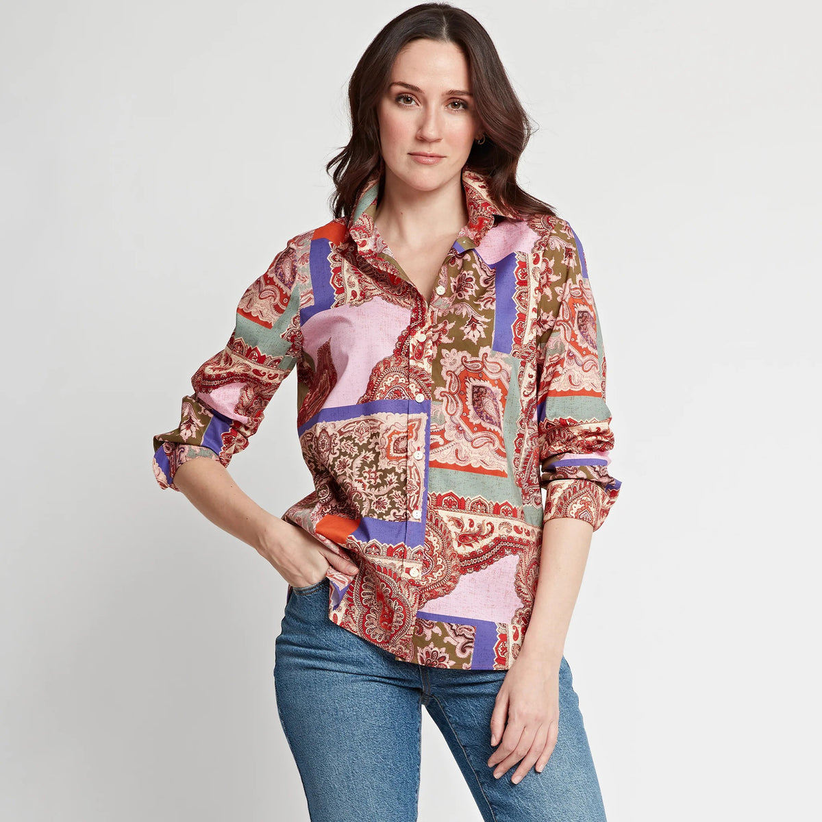 A woman stands against a plain background, wearing a colorful, patterned button-up shirt with pink, red, purple, and green designs, and blue jeans. She has long brown hair and a neutral expression, with one hand in her pocket.