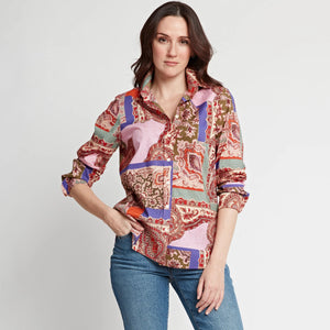 A woman stands against a plain background wearing the HINSON WU - REESE LONG SLEEVE SHIRT, a relaxed fit patchwork paisley top in pink, red, purple, and green, paired with blue jeans. She looks confidently at the camera with one hand in her pocket.