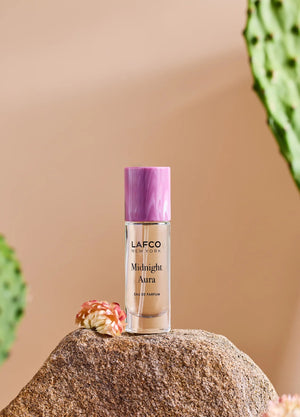 A person holds a 10ml bottle of LAFCO Fine Fragrance, Midnight Aura, on their bare shoulder against a soft purple background.