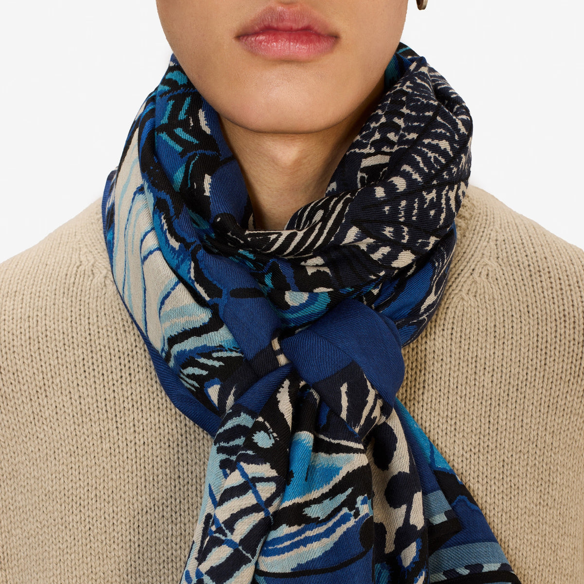 A person wearing a beige knit sweater and an INOUI EDITIONS - PAPILLON (BUTTERFLIES) Wool Scarf in Navy by INOUI, made in India, wrapped around their neck, shown from the chin to the chest.