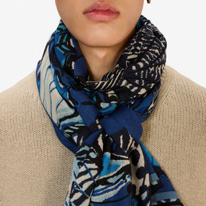 A person wearing a beige knit sweater and an INOUI EDITIONS - PAPILLON (BUTTERFLIES) Wool Scarf in Navy by INOUI, made in India, wrapped around their neck, shown from the chin to the chest.