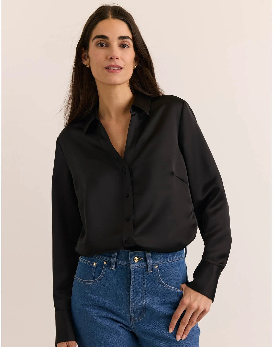 A woman with long dark hair wears the HINSON WU Lindsey Long Sleeve Liquid Satin Shirt in Black, paired with blue jeans, standing against a plain light-colored background.