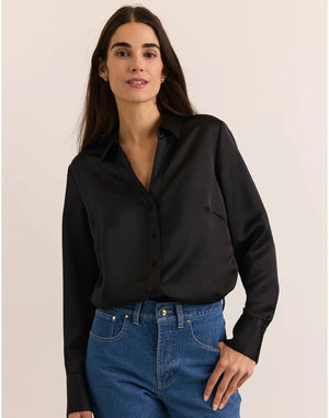 A woman with long dark hair wears the HINSON WU Lindsey Long Sleeve Liquid Satin Shirt in Black, paired with blue jeans, standing against a plain light-colored background.