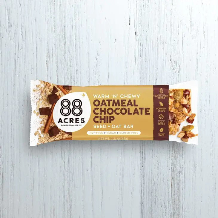 A packaged snack bar labeled &quot;88 ACRES - OATMEAL CHOCOLATE CHIP SEED OAT BAR&quot; from the brand 88 ACRES is shown against a light wooden background. The packaging highlights that it is a seed + oat bar, emphasizing its nut-free, vegan, and gluten-free qualities, and promises creamy chocolate chips for a delightful chewy texture.