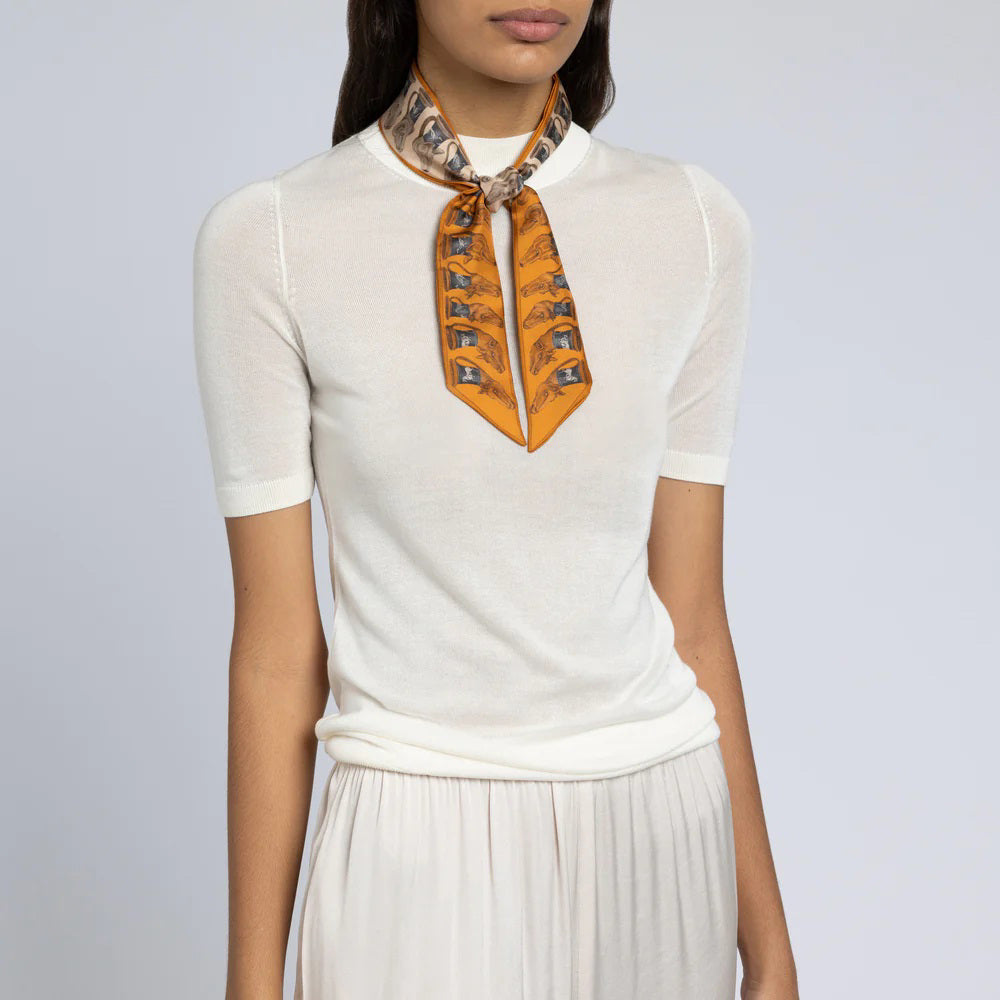 A person is wearing a white short-sleeved top paired with a pleated white skirt. They have SABINA SAVAGE ENGLAND's THE TEMPLE OF PEGASUS SILK RIBBON scarf, which features an orange and gray pattern reminiscent of ancient Greek art, tied neatly around their neck. The background is plain and neutral-colored.