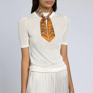 A person is wearing a white short-sleeved top paired with a pleated white skirt. They have SABINA SAVAGE ENGLAND's THE TEMPLE OF PEGASUS SILK RIBBON scarf, which features an orange and gray pattern reminiscent of ancient Greek art, tied neatly around their neck. The background is plain and neutral-colored.