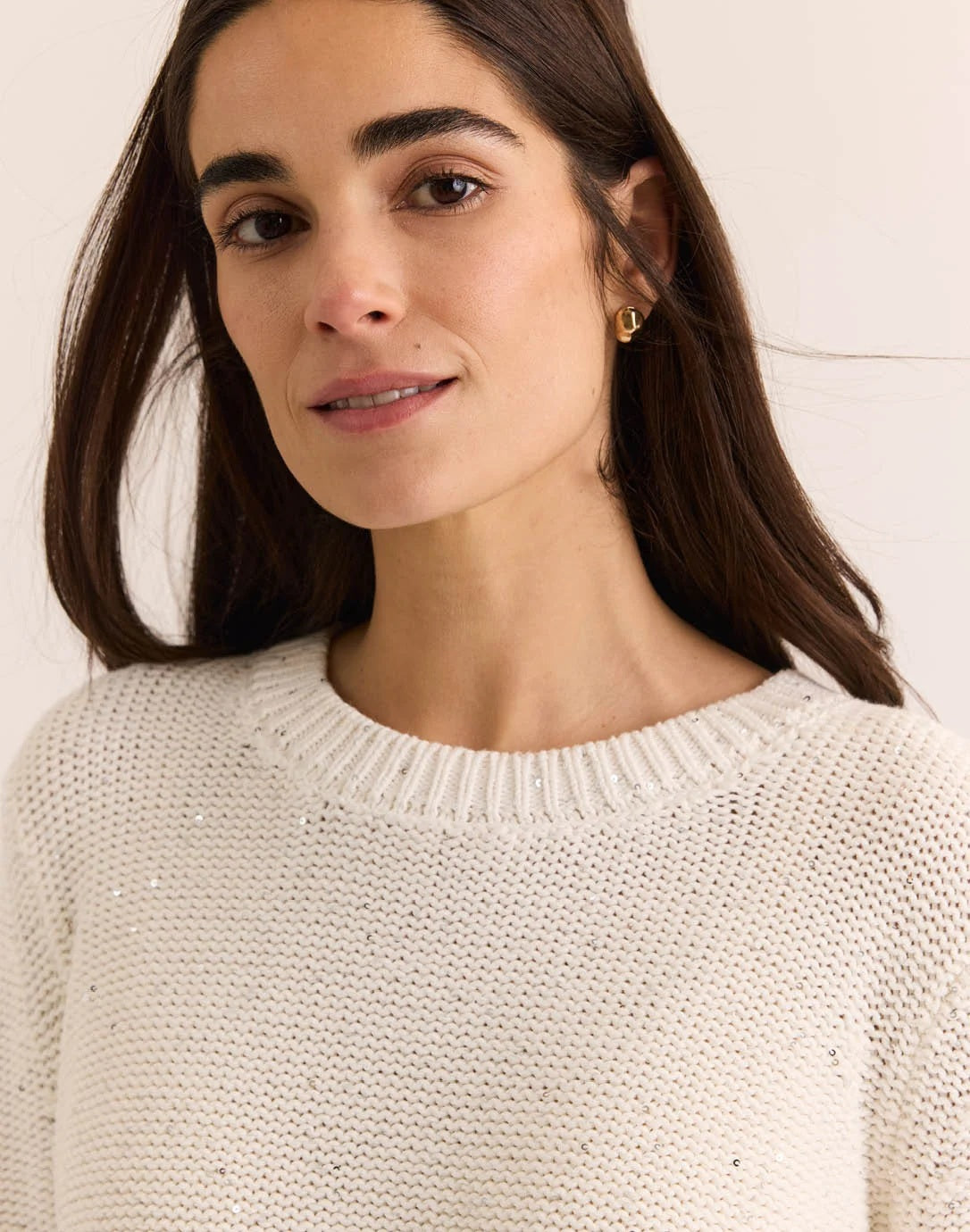 A woman with long brown hair wears the HINSON WU - QUEENIE Long Sleeve Sequin Sweater in Pearl White and blue jeans, standing against a plain light background and looking slightly to the side with a relaxed expression.