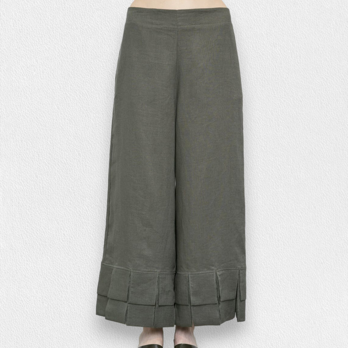 A person is wearing MEIMEIJ&#39;s olive green linen pull-on pants with a pleated bottom design and ruffle cuffs. The pants reach down to their ankles, and the background is a textured white wall. Only the lower half of the body is visible.