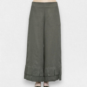 A person is wearing MEIMEIJ's olive green linen pull-on pants with a pleated bottom design and ruffle cuffs. The pants reach down to their ankles, and the background is a textured white wall. Only the lower half of the body is visible.
