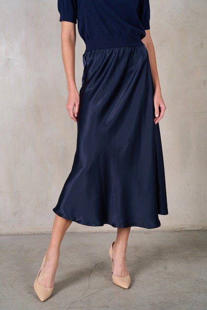 A woman models the ROSE &amp; CROWN - SATIN MIDI SKIRT in silky blue, paired with a short-sleeved light blue knit top, standing against a white background. Her head is not visible in the image.