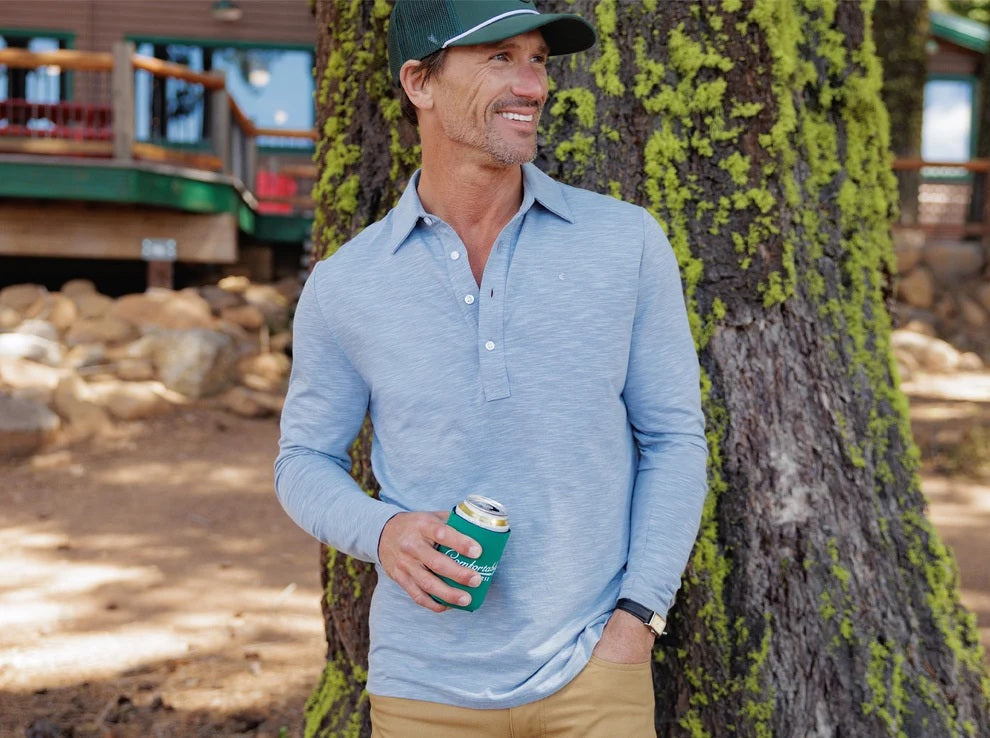 A man with brown hair and a beard is smiling, wearing the CRIQUET - BRRR° Long Sleeve Range Polo in Bluestone and beige pants, with one hand in his pocket, standing against a white background.