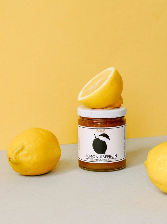 A jar labeled “BRINS - LEMON SAFFRON MARMALADE” from BRINS, garnished with a lemon slice, sits against a light beige background. The jar holds a vibrant yellow marmalade crafted from whole fresh lemons and infused with aromatic saffron for a bright, floral flavor.