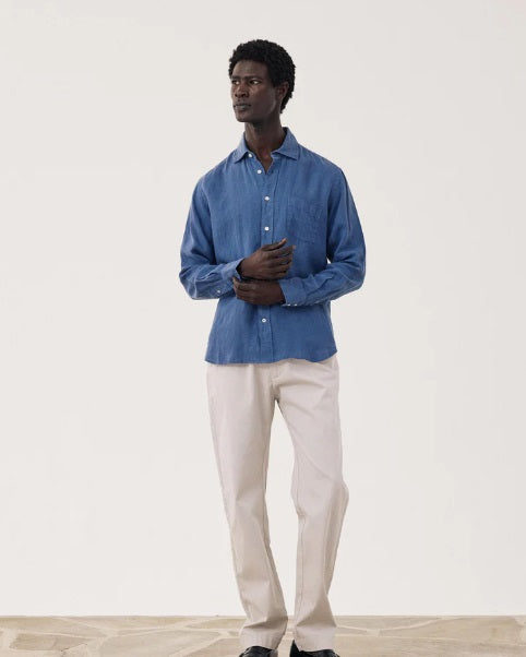 A man with short curly hair poses against a light background, wearing the HARTFORD - PAUL LINEN SHIRT IN DENIM made from European flax linen, featuring a chest pocket, paired with light pants. His right arm is bent behind his back.