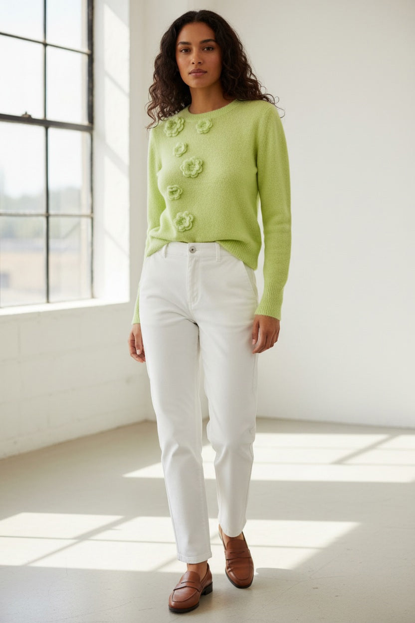 A woman with light brown hair wears the ANNECLAIRE hand embroidered crew neck wool sweater in light green with flower details and a white skirt, standing against a pale background. The timeless piece is handmade in Italy.