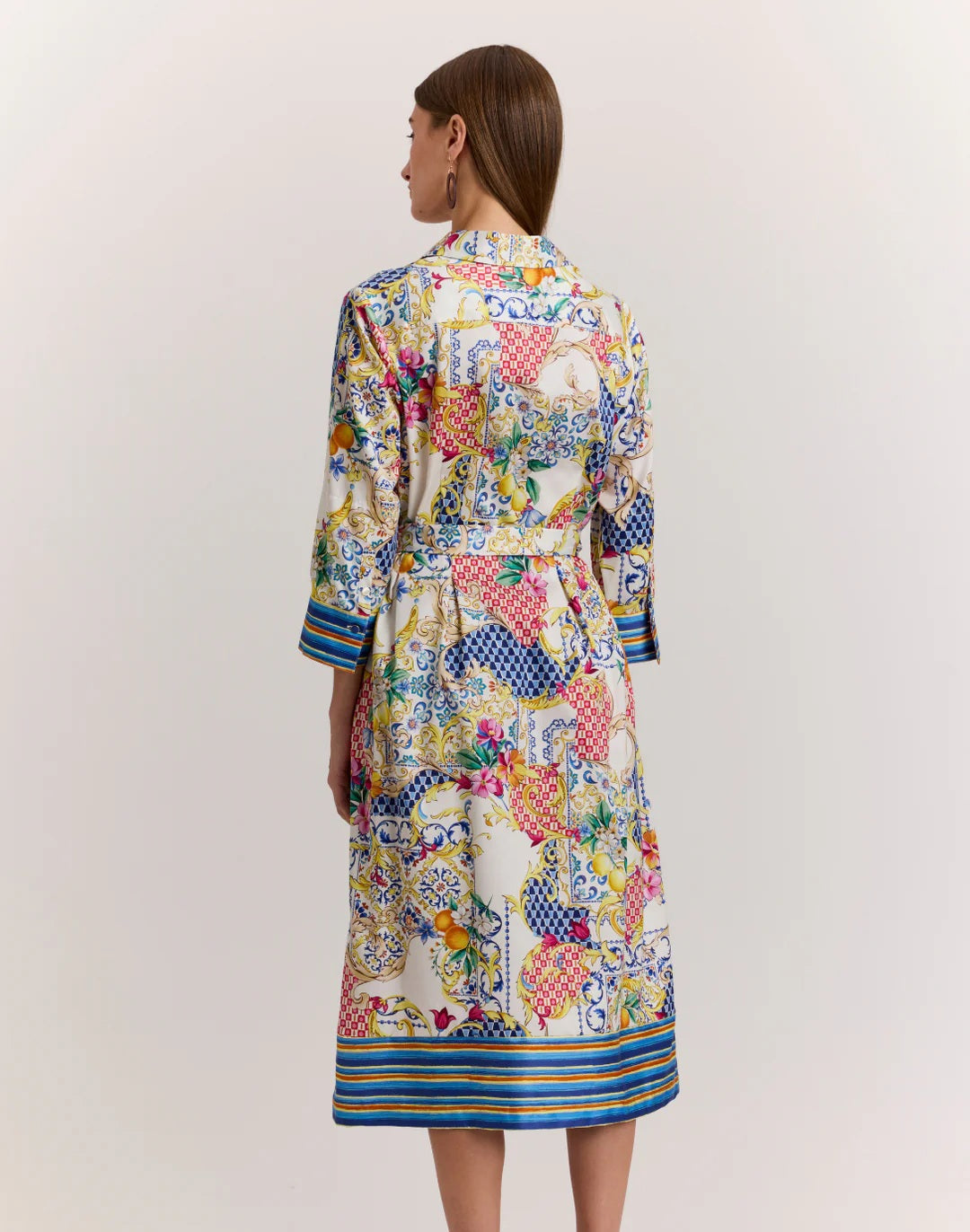 A woman in the HINSON WU Robin 3/4 Sleeve Dress in Amalfi Print—featuring vibrant blue, yellow, red, and white floral and geometric patterns—stands against a plain background.