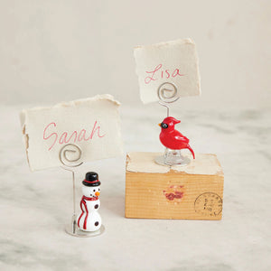 The CREATIVE COOP HANDBLOWN GLASS SNOWMAN PLACECARD HOLDER features a small snowman with a black top hat, red scarf, and black buttons, topped with a spiral metal photo holder.