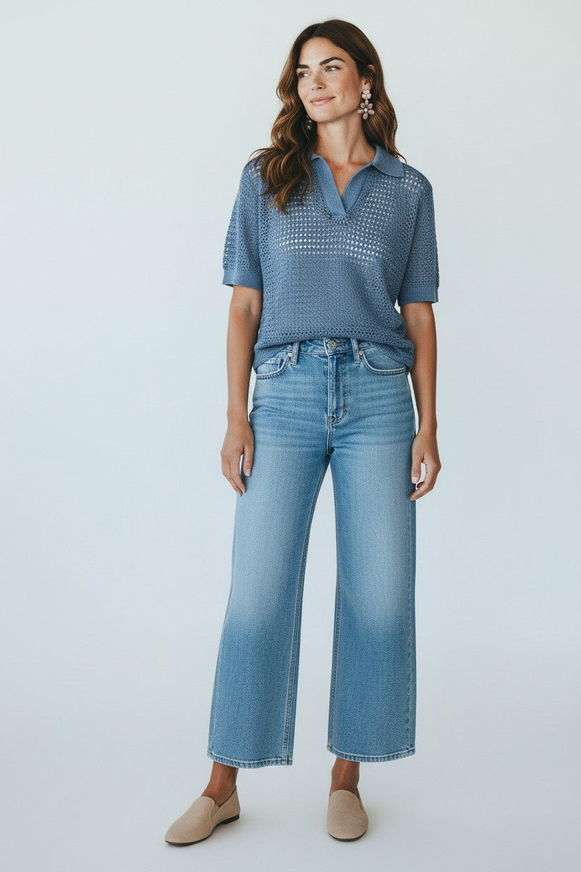 A woman with long brown hair stands against a plain backdrop, smiling slightly as she wears the BASE MILANO - SHORT SLEEVE SPLITNECK COTTON POLO IN LIGHT BLUE by BASE MILANO, paired with light blue wide-leg jeans, beige flats, and statement earrings.