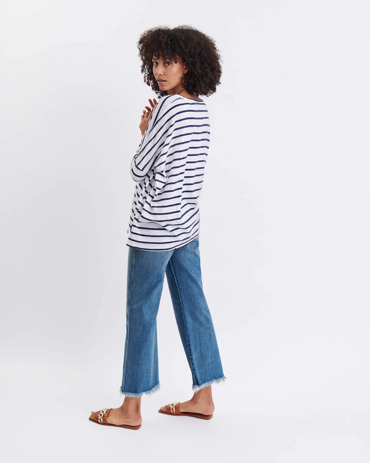 A woman with wavy brown hair models the MERSEA - CATALINA SLUB TEE—an oversized, long-sleeved white top with thin gray stripes—by MERSEA. She pairs it with blue jeans and stands against a plain, light background, hands behind her back.