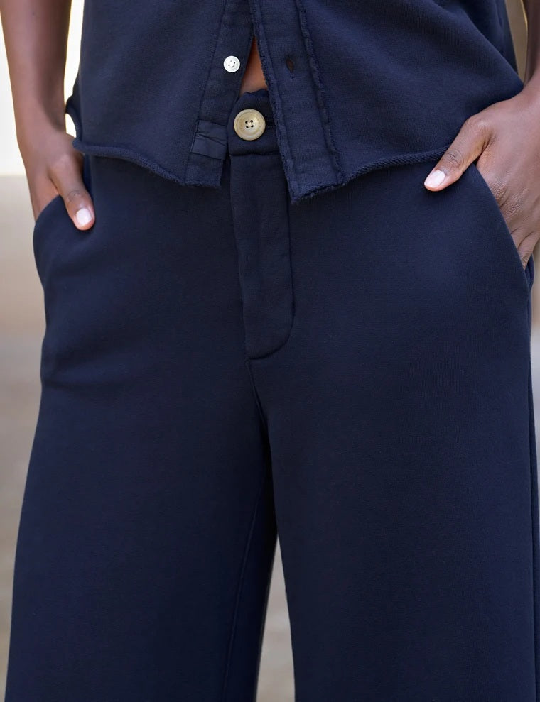 A person stands outdoors facing away, wearing the FRANK & EILEEN Riley Trouser Sweatpant in Triple Fleece Classic Navy paired with black shoes, one hand resting by their side near a wooden wall and a bench—a relaxed look inspired by FRANK & EILEEN’s California Heritage Collection.