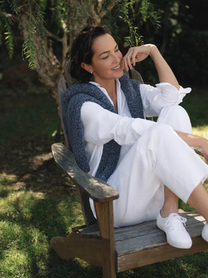 A person with wavy blonde hair, wearing a relaxed fit FRANK & EILEEN Montecito Crew Neck Cardigan in Pure Italian Cotton Blueberry and white pants, stands outdoors facing away, surrounded by greenery and trees.