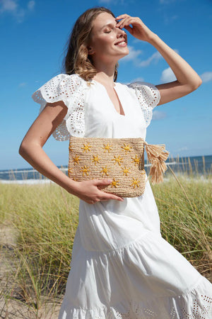 The MAR Y SOL - SOLEIL CLUTCH by MAR Y SOL, featuring yellow sun embroidery and an oversized tassel, rests on a light cloth with three lemons and brown sunglasses. Sunlight casts playful shadows across this stylish display.