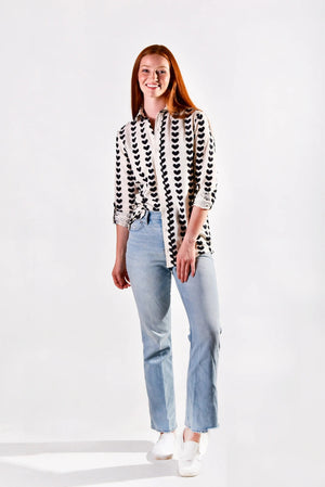 A person wearing the EMILY PHILLIPS - BLACK HEARTS GIRLFRIEND BUTTON DOWN SHIRT, a sustainably made white cotton top featuring rolled sleeves and vertical rows of large black heart patterns.
