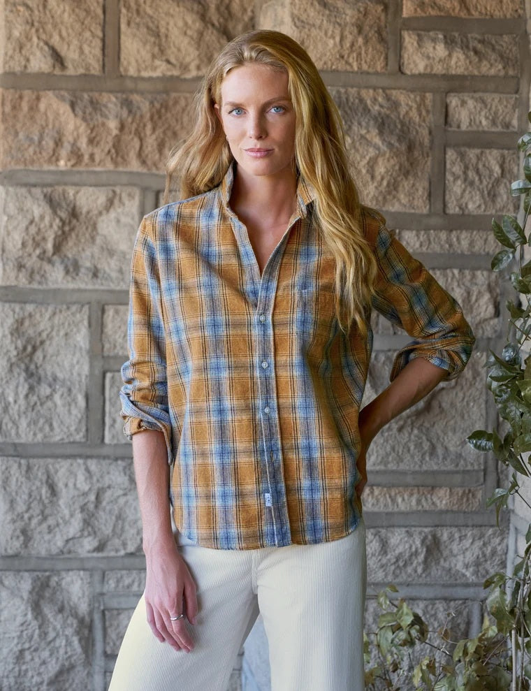 A woman stands indoors against a neutral background with soft light streaming in, smiling gently with one hand in her pocket. She wears the FRANK & EILEEN Eileen Relaxed Button Up Shirt in Italian Flannel Gold Blue Large Plaid—featuring a bust-flattering button placement—paired with cream high-waisted pants and brown ankle boots.