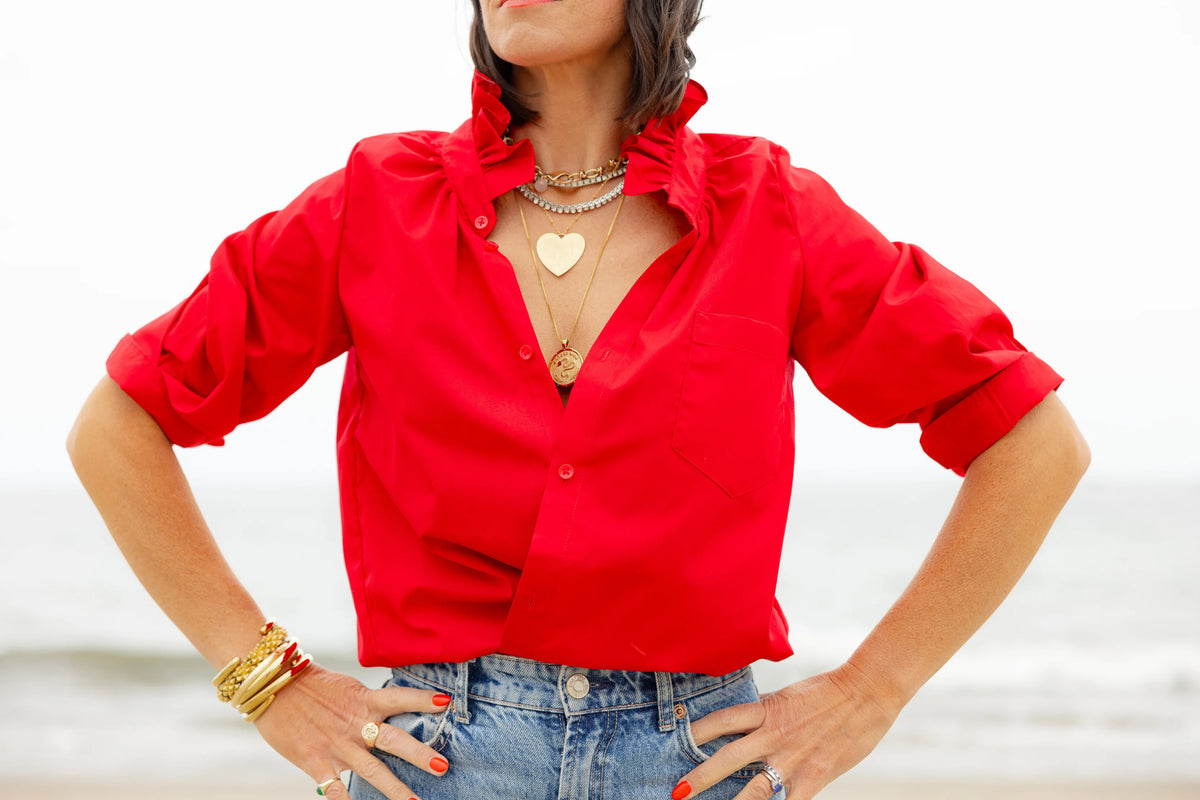 A woman in the KMJ - Southern Lover Shirt with a ruffle collar and blue jeans, featuring the iconic KMJ tuck, stands confidently beside a horse, holding its reins amidst green foliage. She's elegantly posed with jewelry, embodying Southern-inspired charm.
