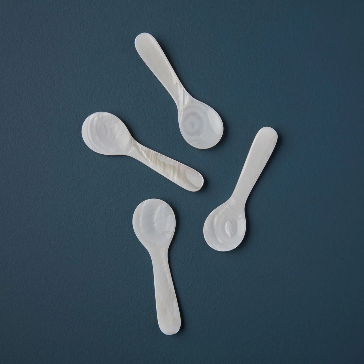 Four BE HOME - SEA SHELL SPOON SINGLE spoons by BE HOME, INC are arranged in a scattered pattern on a dark blue surface.