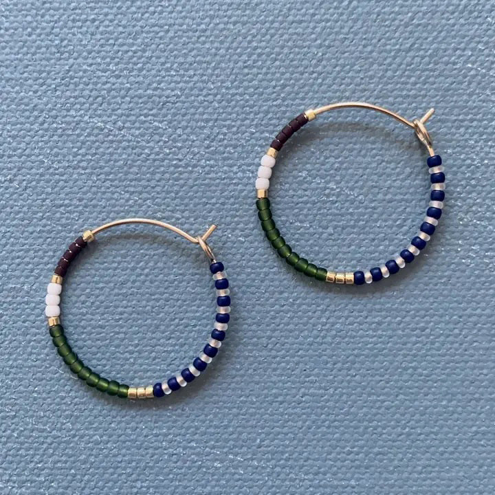 The ALICE RISE - MIDI COLORLOOP EARRINGS IN OLIVE by ALICE RISE feature 14k gold filled hoops adorned with white, navy blue, green, burgundy, and gold Japanese glass beads, displayed on a textured light blue surface.