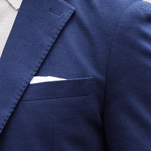 A man wearing sunglasses, the COLLARS AND CO MAVERICK KNIT UNSTRUCTURED BLAZER IN NAVY over a white shirt, light pants, and brown loafers stands on a sidewalk in front of a light stone building.