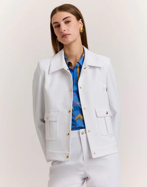 A woman with straight brown hair wears the HINSON WU - HANNAH TRAVELER'S TWILL JACKET with gold buttons over a blue patterned shirt and white pants, standing against a plain light background.