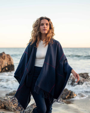 A woman with wavy brown hair wears the MERSEA Plaid Pocket Wrap—a black, white, and red plaid poncho with fringed edges—over a white outfit indoors. This MERSEA travel wrap is perfect for cozy style on the go.