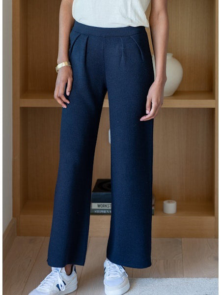 Seated barefoot on a light beige chair, a person wears MERSEA’s SEAPORT KNIT PULL ON PANT with side pockets, paired with a white top and open shirt. One hand rests in their pocket as sunlight streams onto their legs.