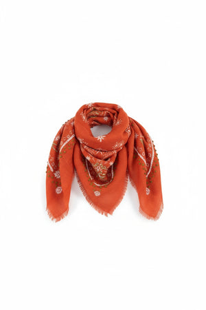 The STORIATIPIC - NELLY WOOL BANDANA SCARF by STORIATIPIC is an orange wool accessory measuring 100x100 cm, featuring fringed edges and adorned with white and pink floral embroidery accented with green details. Elegantly arranged in a loose loop on a white background, this scarf offers both style and comfort.