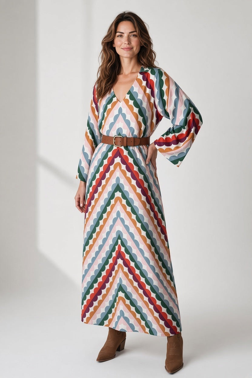 A woman wears a BRITON COURT MILANI MAXI DRESS IN GROOVY GROVE, a long-sleeved, v-neck maxi dress with a multicolored scalloped chevron print and brown ankle boots.
