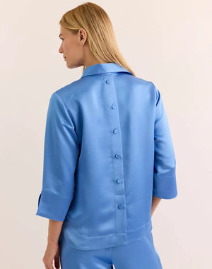 A woman with blonde hair wears the HINSON WU Aileen 3/4 Sleeve Liquid Satin Shirt in Sky Blue—a loose-fitting blouse featuring an elegant drape and luminous sheen. Paired with matching pants, the light blue satin fabric from HINSON WU catches the light beautifully as she stands against a simple, light background.
