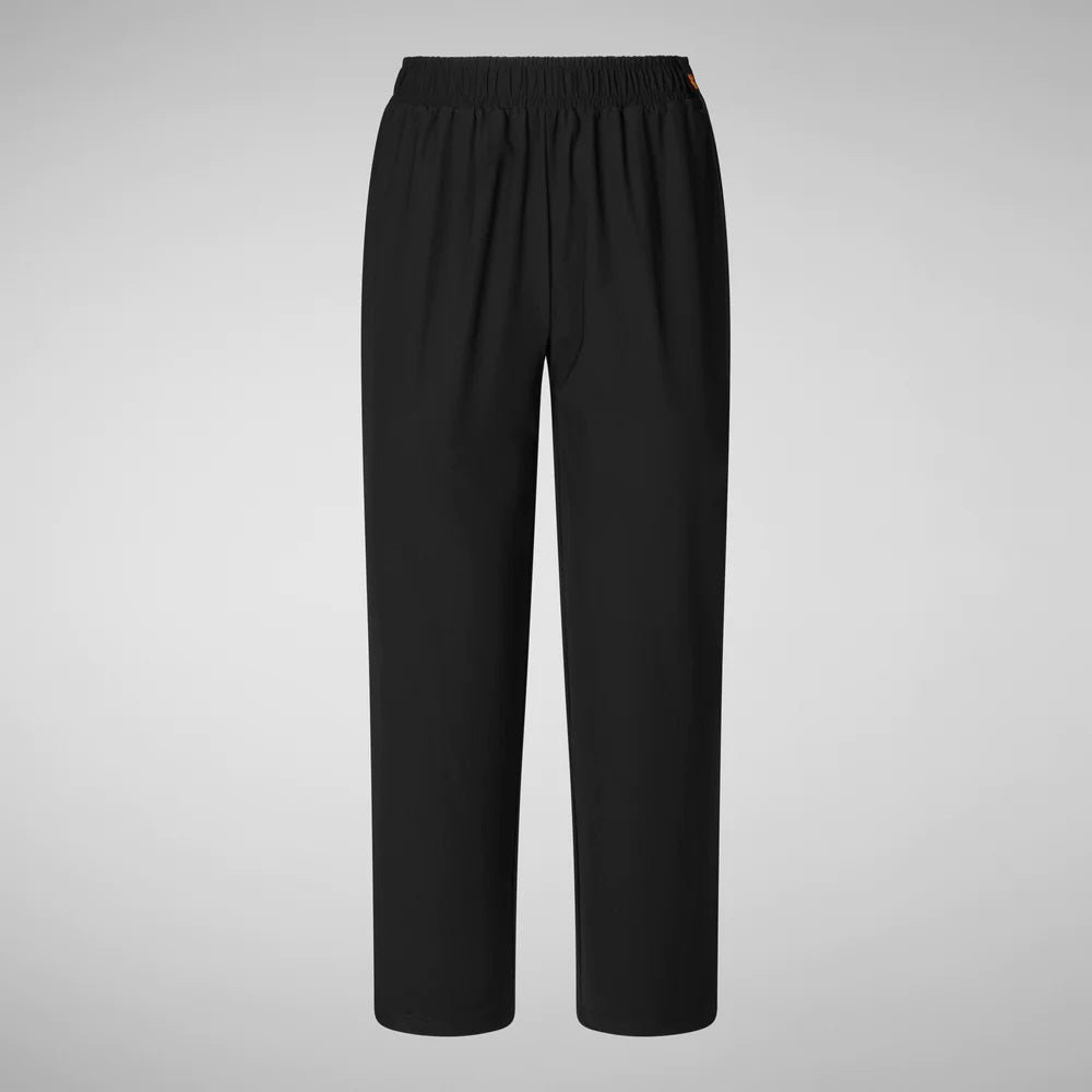 A person with shoulder-length hair and a neutral expression wears a white long-sleeve shirt, SAVE THE DUCK MILAN PULL ON PANT in black wrinkle-free fabric, and black slip-on shoes. Their hands rest at their sides against a plain background.