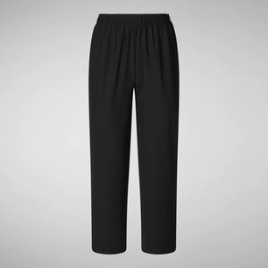 A person with shoulder-length hair and a neutral expression wears a white long-sleeve shirt, SAVE THE DUCK MILAN PULL ON PANT in black wrinkle-free fabric, and black slip-on shoes. Their hands rest at their sides against a plain background.