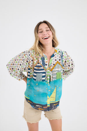 A smiling woman stands with her hands in her pockets, wearing the STORIATIPIC COCO GERTRUDE BLOUSE—featuring polka dot sleeves and a beach hut print—paired with beige shorts, against a plain white background.