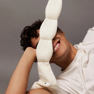 The NODPOD Weighted Sleep Eye Mask in Bone by NODPOD is a beige, segmented, plush mask with wider ends and a gently wavy, narrower middle section, shown laid flat on a white background—ideal for providing gentle pressure and soothing comfort as a weighted eye mask.