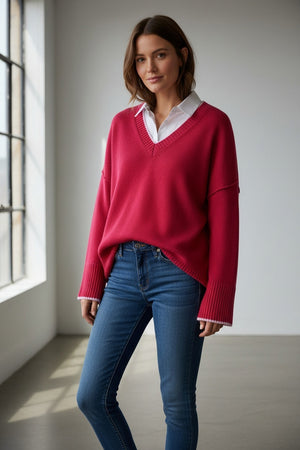Wearing the MERSEA Montauk Colorblock V Neck Sweater in Raspberry, a woman in jeans poses against a pink background, smiling softly and raising one arm near her face.