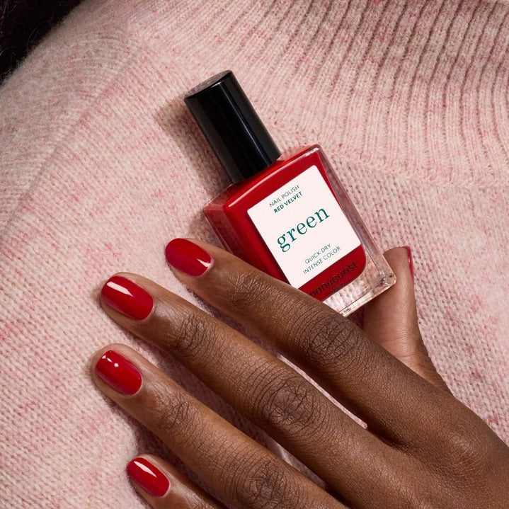 A hand with neatly manicured nails painted in MANUCURIST - GREEN™ NAIL POLISH IN RED VELVET by MANUCURIST INC rests on a textured red leather surface, creating a coordinated and stylish look.